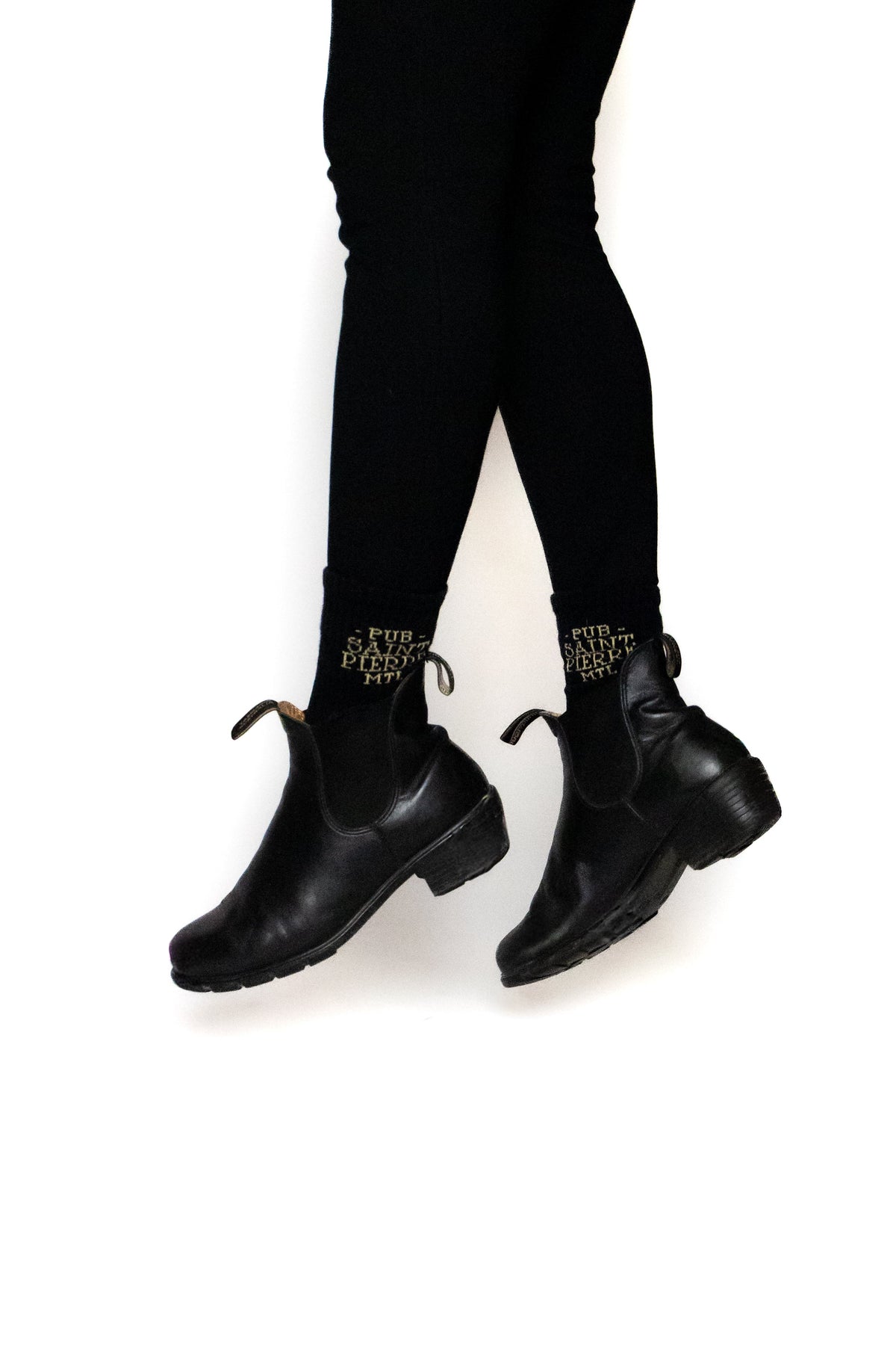 Mid-air in black boots and Pub Saint Pierre crew socks, gold lettering visible at the ankle