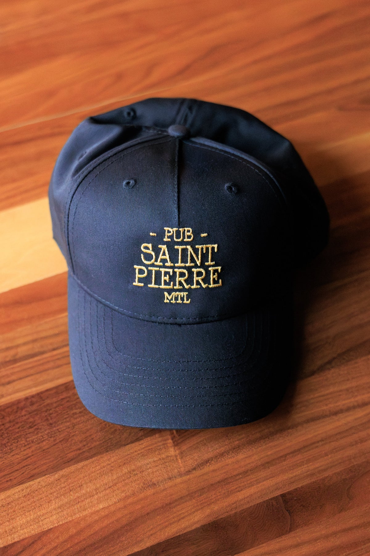 Navy Pub Saint Pierre baseball cap with gold embroidered wordmark, laid on a walnut table