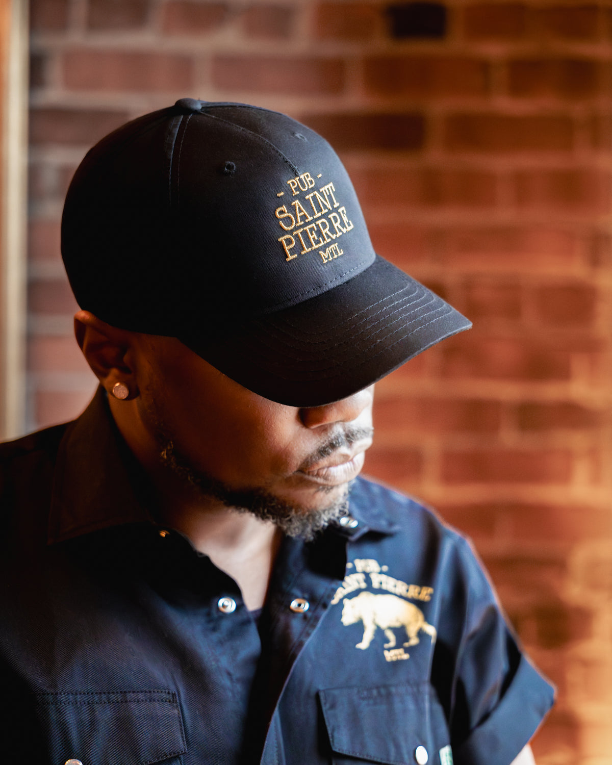 Wearing the navy Pub Saint Pierre baseball cap in profile against a brick wall, gold embroidered wordmark