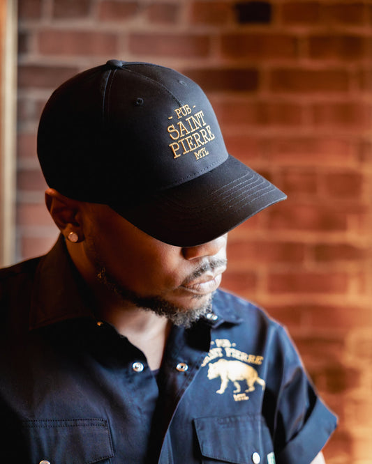 Wearing the navy Pub Saint Pierre baseball cap in profile against a brick wall, gold embroidered wordmark