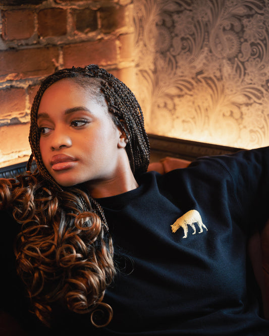 Seated against exposed brick wearing the black Pub Saint Pierre crewneck sweatshirt, gold bear at the chest