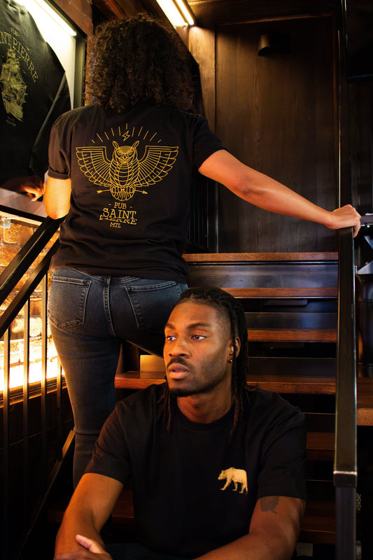Two people on the pub stairs in black Pub Saint Pierre tees, gold owl on the back and gold bear at the chest