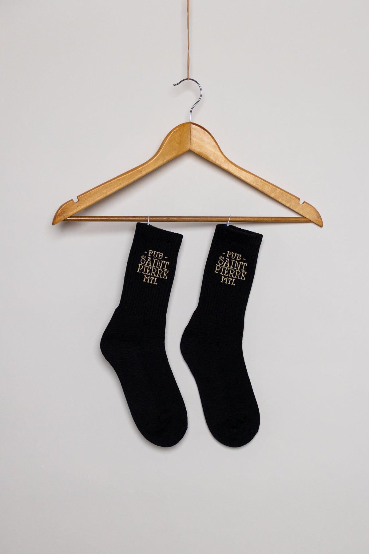 Black Pub Saint Pierre crew socks on a hanger against a plain background, gold PUB SAINT PIERRE MTL lettering at the cuff