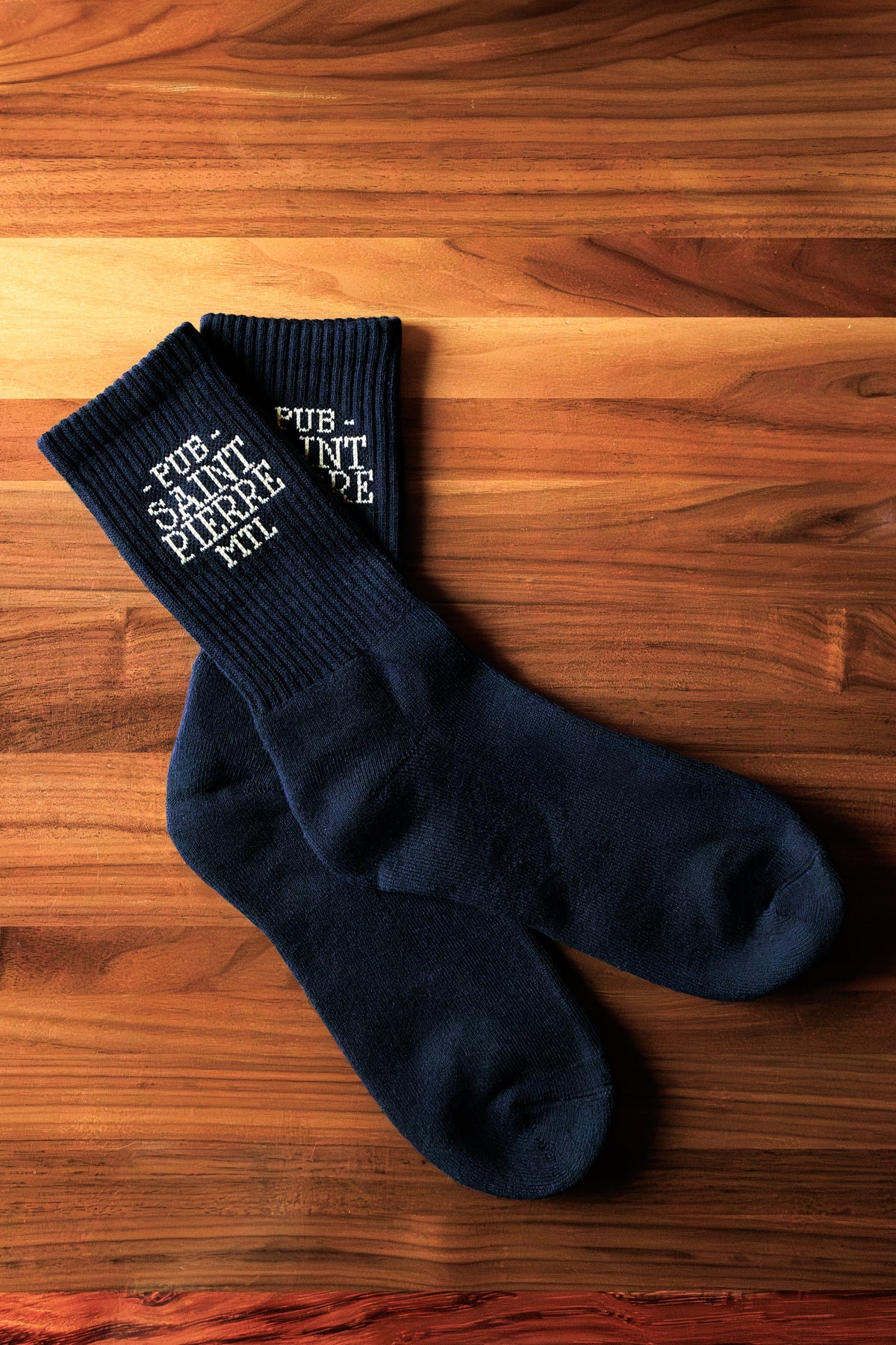 A pair of black Pub Saint Pierre crew socks with gold lettering, laid on a walnut table