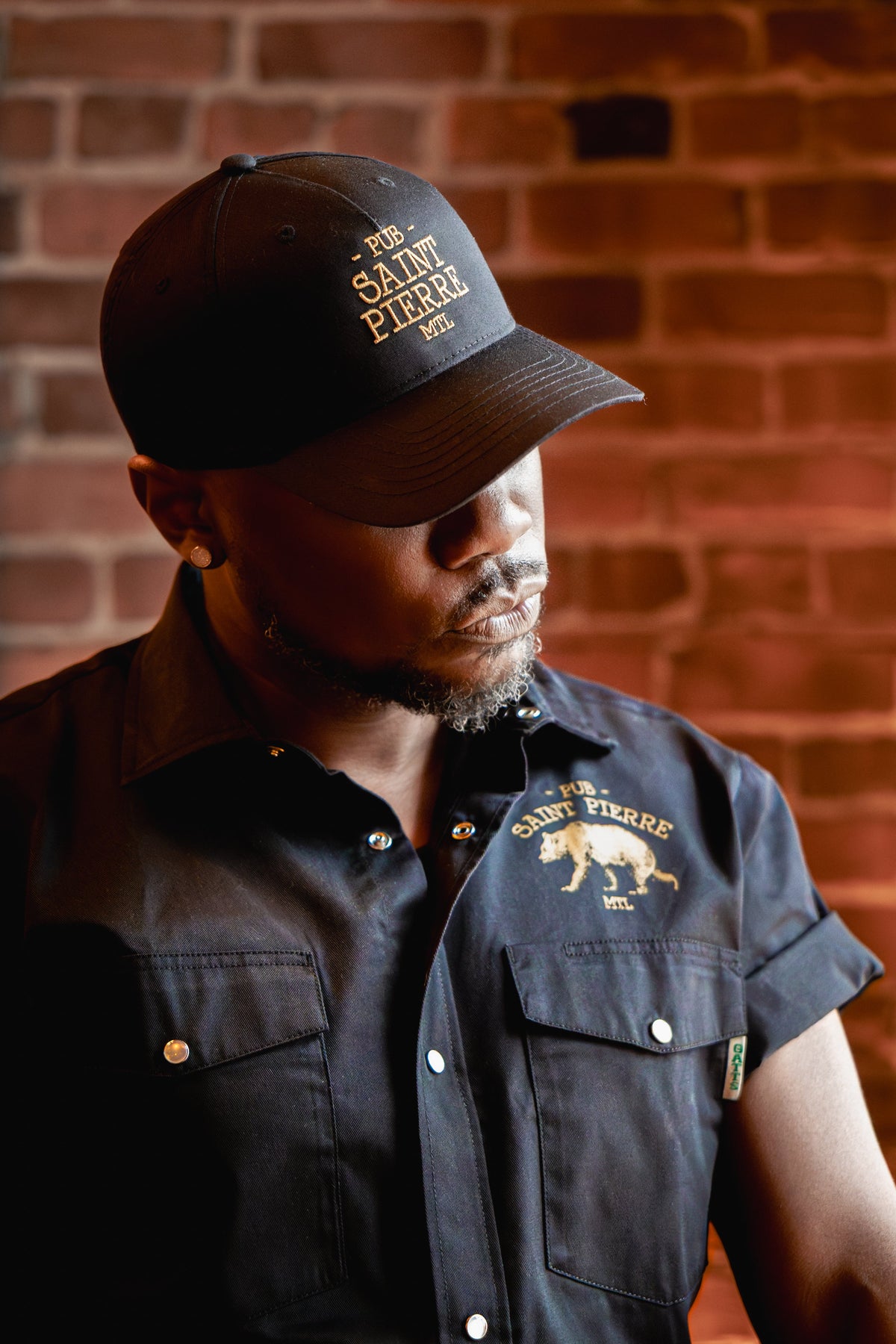Man wearing the Pub Saint Pierre work shirt and cap against a brick wall, gold bear at the chest